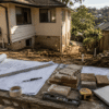 Architectural plans, tools and material samples on a weathered workbench overlooking an Australian suburban renovation site.